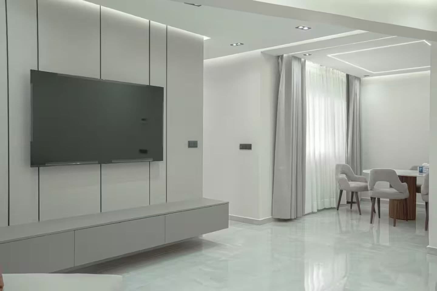 Un salon moderne doté d’un téléviseur à écran plat fixé au mur, d’un meuble de rangement intégré et d’un coin repas avec une table et des chaises.