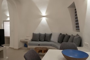A modern living room with a curved sofa, a coffee table, and a television. - Santoterra Antigua Luxury Suites (Santorini)