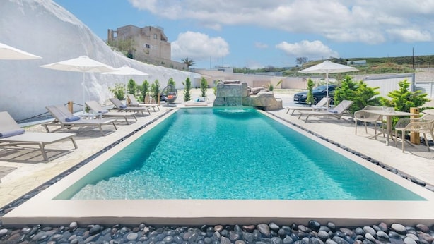 Una piscina rectangular con tumbonas y sombrillas, rodeada por un borde rocoso y un castillo al fondo.