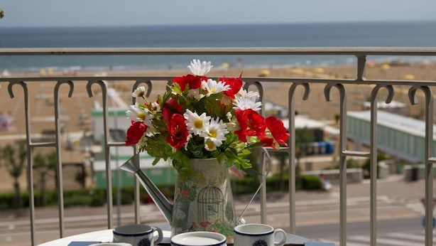 Mesa de balcón con bandeja, un jarrón de flores y tazas a juego, con vistas a la playa y al paisaje urbano.