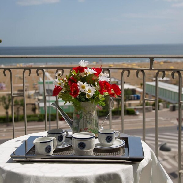 A balcony table with a tray, a pitcher of flowers, and matching cups, overlooking a beach and cityscape. - Hotel Ancora (Rimini)