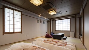 Ein traditioneller japanischer Raum mit Tatami-Boden, zwei Betten mit gemusterten Bettbezügen, einem kleinen Tisch und einem Fernseher.
