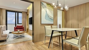 Ein modernes Hotelzimmer mit einem großen Fenster, einer Essecke mit rundem Tisch und Stühlen, einem roten Sofa und einem großen abstrakten Gemälde an der Wand.