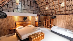 Habitación con una cama grande, una cama más pequeña y un banco de madera. Se observa un armario con espejo y repisas. La habitación tiene techo de paja y paredes de madera.
