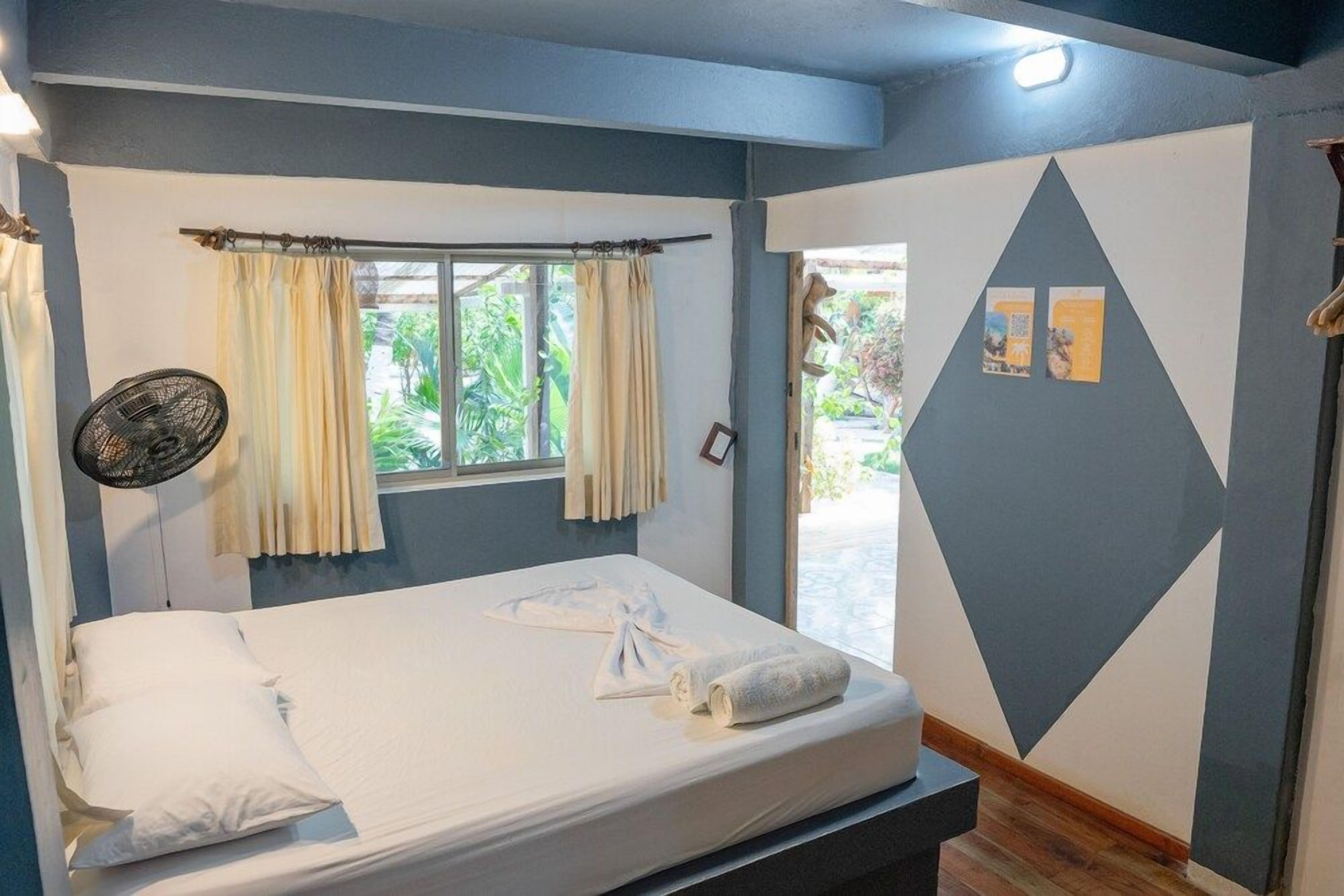 Una cama individual con colchón blanco, dos almohadas y una toalla enrollada. Se ve una ventana con cortinas, un ventilador de pared y una puerta que da al exterior.