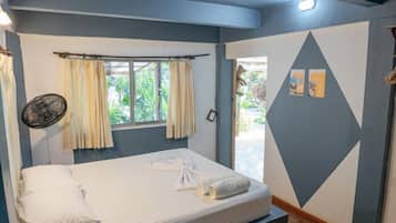 Una cama individual con colchón blanco, dos almohadas y una toalla enrollada. Se ve una ventana con cortinas, un ventilador de pared y una puerta que da al exterior.