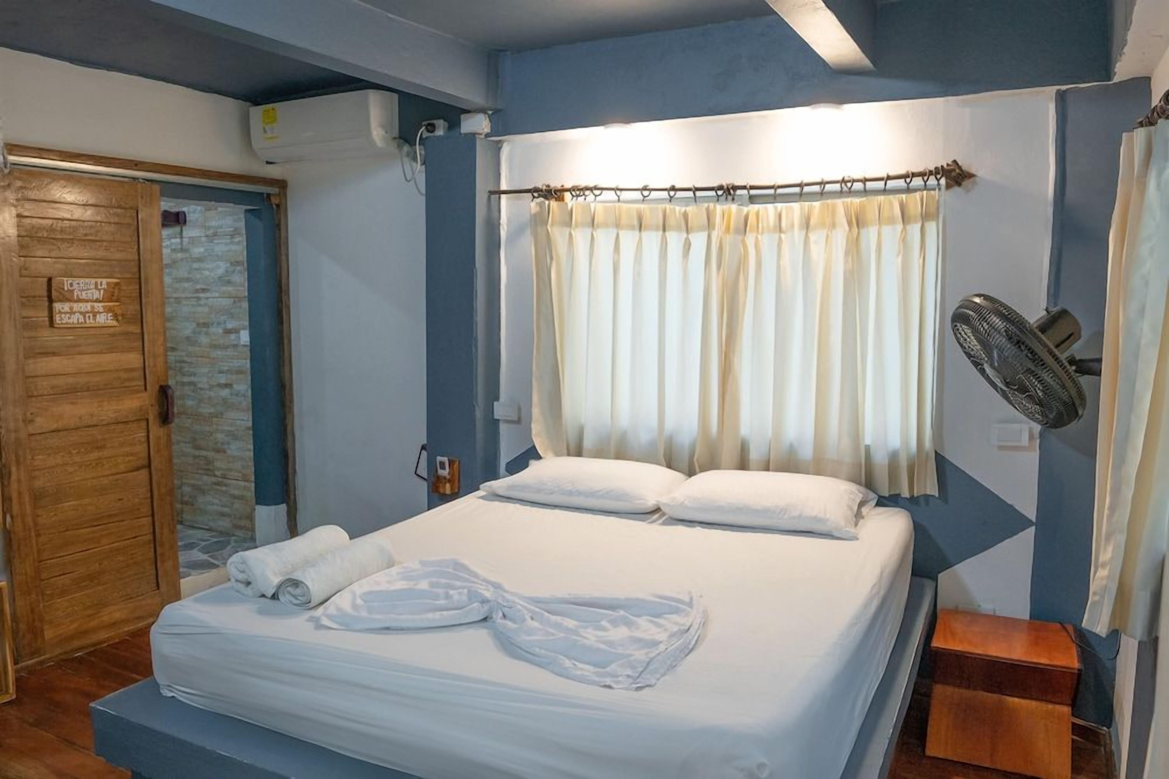 A hotel room with a bed, a wooden door, a curtain, a fan, and a sign on the wall.