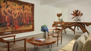 A room with a large traditional painting, a wooden coffee table with books, a vase with flowers, and a unique wooden chair.