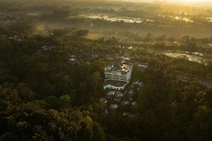 A large, multi-story hotel with a central courtyard, surrounded by dense greenery and agricultural fields.