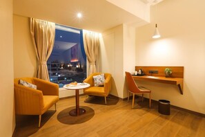 A hotel room with a view of a cityscape at night, two armchairs, a small round table, a wooden side table with a plant, and a hanging light fixture. - IRA by Orchid Sambhajinagar (Chhatrapati Sambhajinagar)