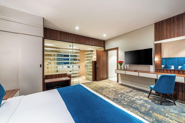 A hotel room with a bed, a desk with a flat-screen TV, a wardrobe, and a glass-enclosed bathroom.