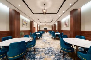 A hotel conference room with round tables and blue chairs, wooden paneling, and a patterned carpet.