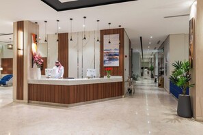 Zona de recepción de un hotel con un mostrador de madera, una recepcionista y un pasillo con plantas en macetas.