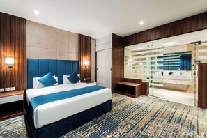 Habitación de hotel moderna con una cama grande, un baño con paredes de vidrio y una pared decorativa de madera.