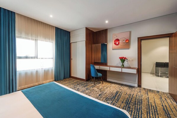 A hotel room with a bed, a desk, a chair, a desk lamp, a vase with flowers, and a window with curtains.