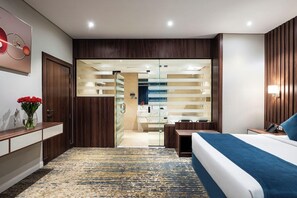 A modern hotel room with a bed, bedside table, and a bathroom visible through a glass door.