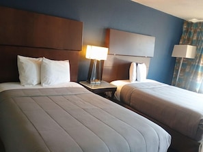 Habitación de hotel con dos camas, una mesita de noche con lámpara y un ventanal con cortinas.