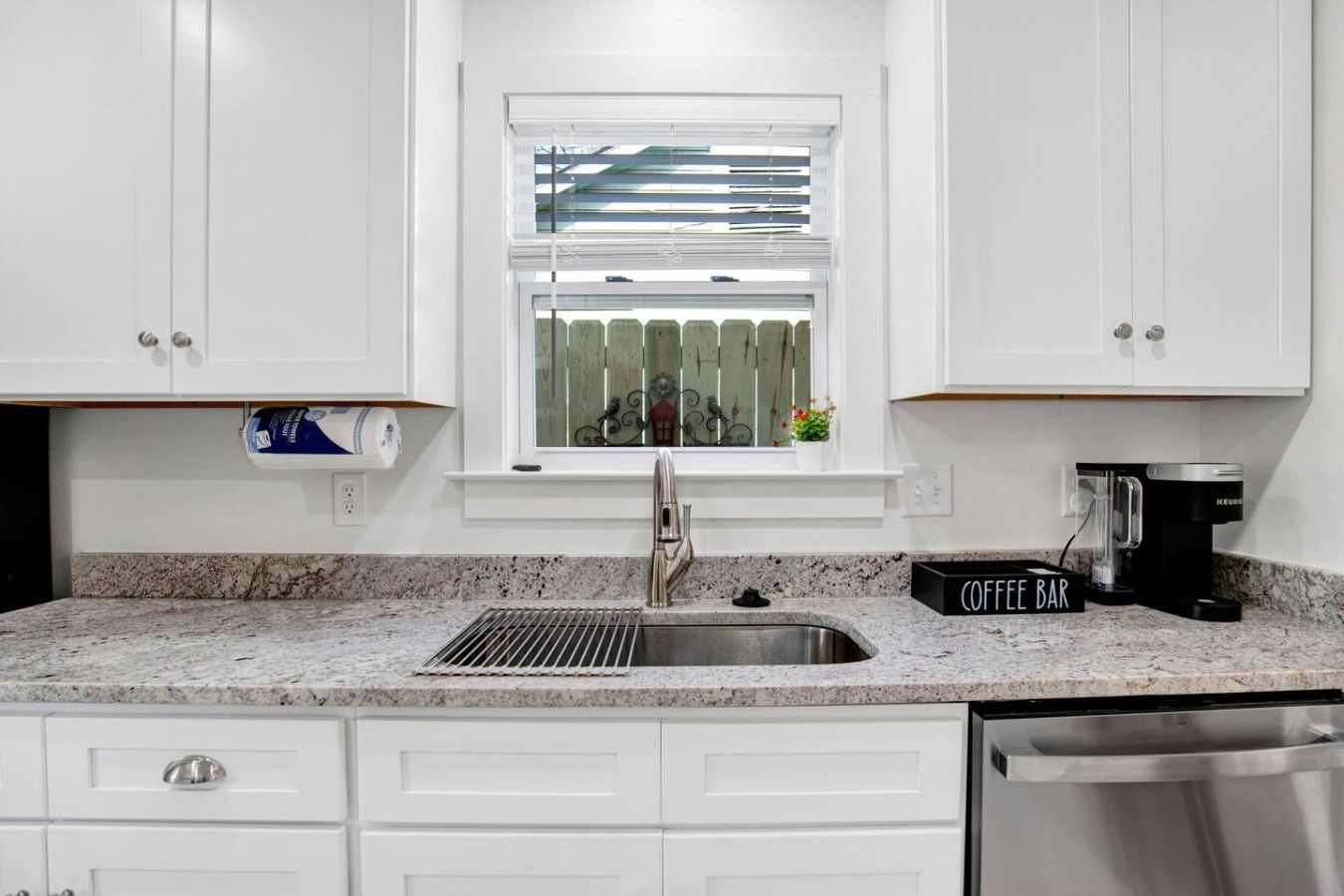 A kitchen with white cabinets, a stainless steel sink, a coffee maker, and a window with blinds.