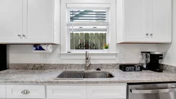A kitchen with white cabinets, a stainless steel sink, a coffee maker, and a window with blinds.