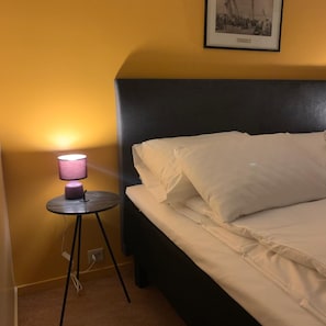 Una cama con sábanas blancas y un cabecero negro, una mesita redonda con una lámpara rosa, y un cuadro colgado en la pared.