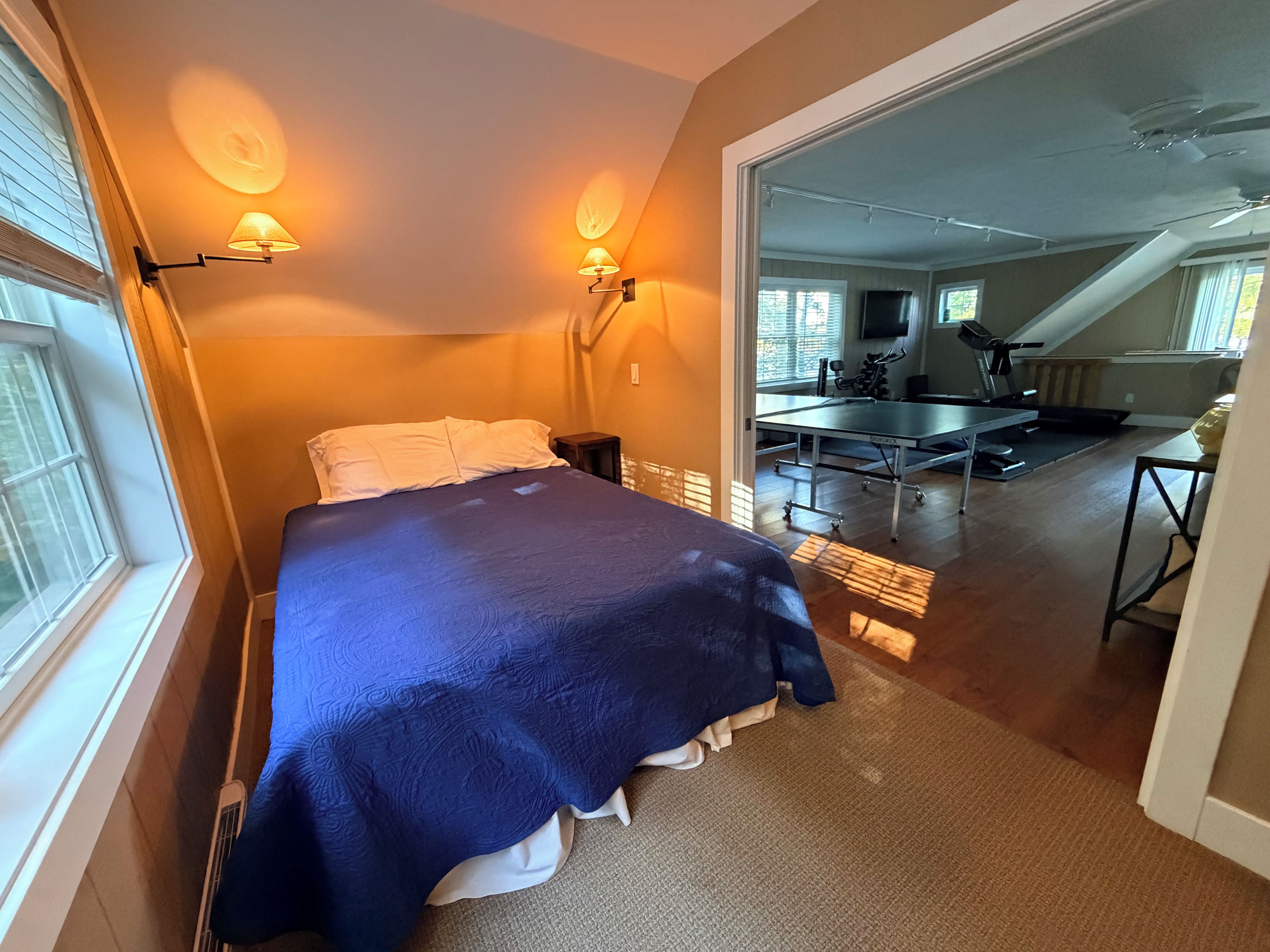 Ein Schlafzimmer mit einem Bett, zwei Wandleuchten, einem Fenster und Blick in einen Wohnbereich mit Laufband und Trainingsgeräten.