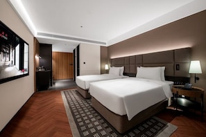 Habitación de hotel con dos camas, un escritorio, una silla, una lámpara y un televisor montado en la pared.
