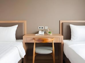 A hotel room with two beds, a wooden desk, a chair, and a bedside table with a lamp and a small plant. - Grand Caman Hotel (Bekasi)