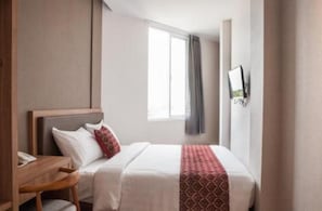 A hotel room with a bed, a desk, a chair, a window with curtains, and a wall-mounted TV. - Grand Caman Hotel (Bekasi)