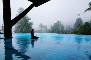 A person meditating in a clear blue pool with a view of palm trees and a mountain in the background. - Grand Kanaya Baturraden (Baturaden)