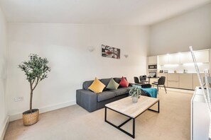 A modern living room with a grey sofa, a coffee table, and a dining area with a kitchen in the background. - Surrey's Stylish 2-Bed | Parking | Near Station - Pass the Keys (Surrey)
