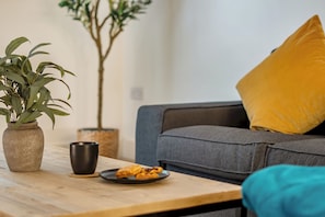 A living room with a sofa, a coffee table, a plant, and a plate of pastries. - Surrey's Stylish 2-Bed | Parking | Near Station - Pass the Keys (Surrey)