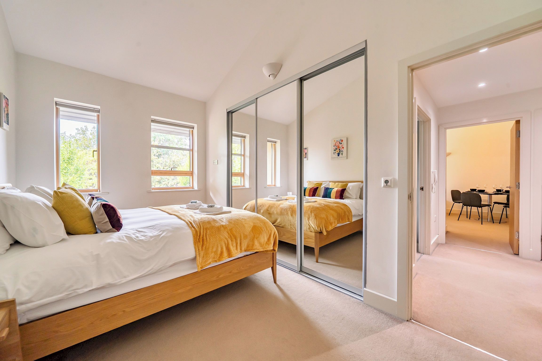 Une chambre moderne avec un grand lit, une porte-fenêtre coulissante et un coin repas visible à travers une porte ouverte.