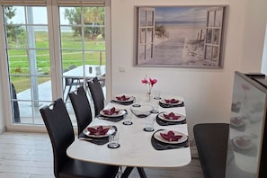 A dining area with a table set for four, chairs, a painting of a beach scene, and a view of a grassy area through large windows. - For 8 guests: entire floor with a garden on the Peenestrom (Kröslin)