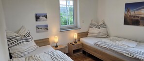 A bedroom with two beds, a nightstand, a lamp, and a window with a view of greenery. - Vrbo Property (Kröslin)