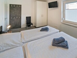 Ein Hotelzimmer mit zwei Betten, einem Schrank, einem Sessel, einem Schreibtisch mit Lampe und einem an der Wand befestigten Fernseher.