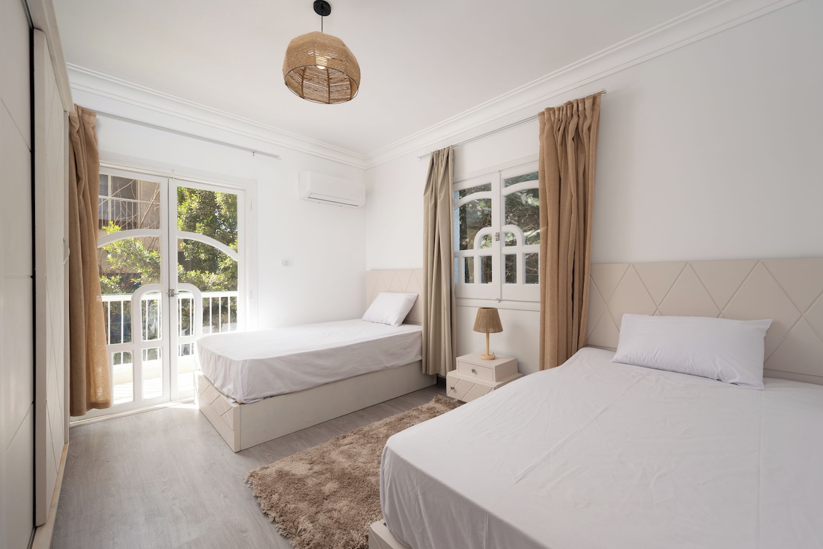 Ein Schlafzimmer mit zwei Betten, einem fensterbelaubten Fenster, einer Deckenleuchte und Blick auf Grünflächen.