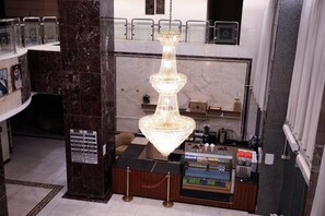 A hotel reception area with a marble column, a chandelier, a counter with a beverage dispenser, and a security camera. - Raafat Al Marzouki Al Kutbi Hotel (macch)