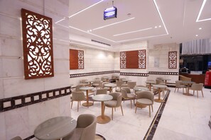A modern interior with round tables and chairs, decorative wall panels, and a kitchen area in the background. - Raafat Al Marzouki Al Kutbi Hotel (macch)