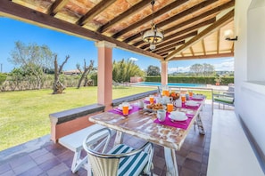 An outdoor patio with a wooden table set for breakfast, surrounded by chairs, and overlooking a swimming pool and garden. - Vrbo Property (Pollenca)
