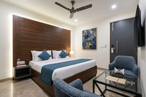 Une chambre d’hôtel avec un lit, une chaise, une table en verre, un ventilateur de plafond, un tableau et un téléviseur.
