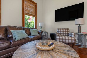 Uma sala de estar com sofá de couro marrom, poltrona xadrez, mesa de centro redonda com um objeto decorativo e televisão fixada na parede.
