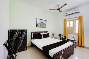 A hotel room with a bed, a desk, a chair, a ceiling fan, an air conditioner, a window with yellow curtains, and a framed picture on the wall. - Hotel O Kelambakkam Bus Terminal (Chennai)