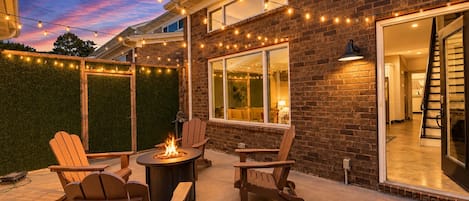 Een terras buiten met een vuurkorf, houten stoelen en sfeerverlichting tegen een bakstenen muur.