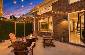 An outdoor patio area with a fire pit, wooden chairs, and string lights against a brick wall. - ✨ Modern 2BR Lyndon Loft Oasis | Work & Play| Private Patio | 2-Car Garage (Louisville)