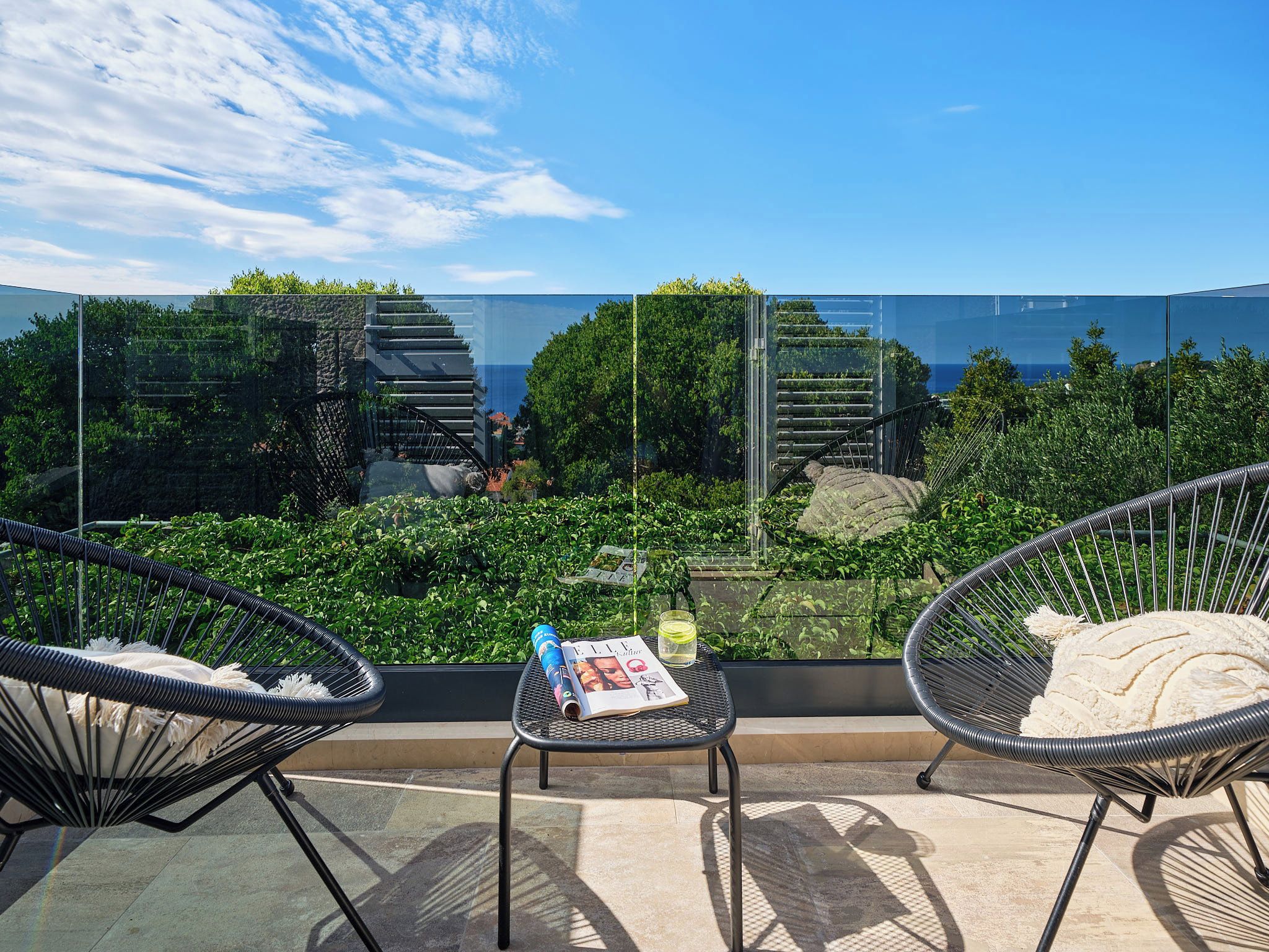 Un balcón con dos sillas de mimbre, una mesita y baranda de cristal con vistas a un jardín y el mar.