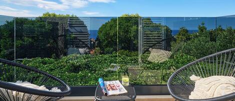 Un balcón con dos sillas de mimbre, una mesita y baranda de cristal con vistas a un jardín y el mar.