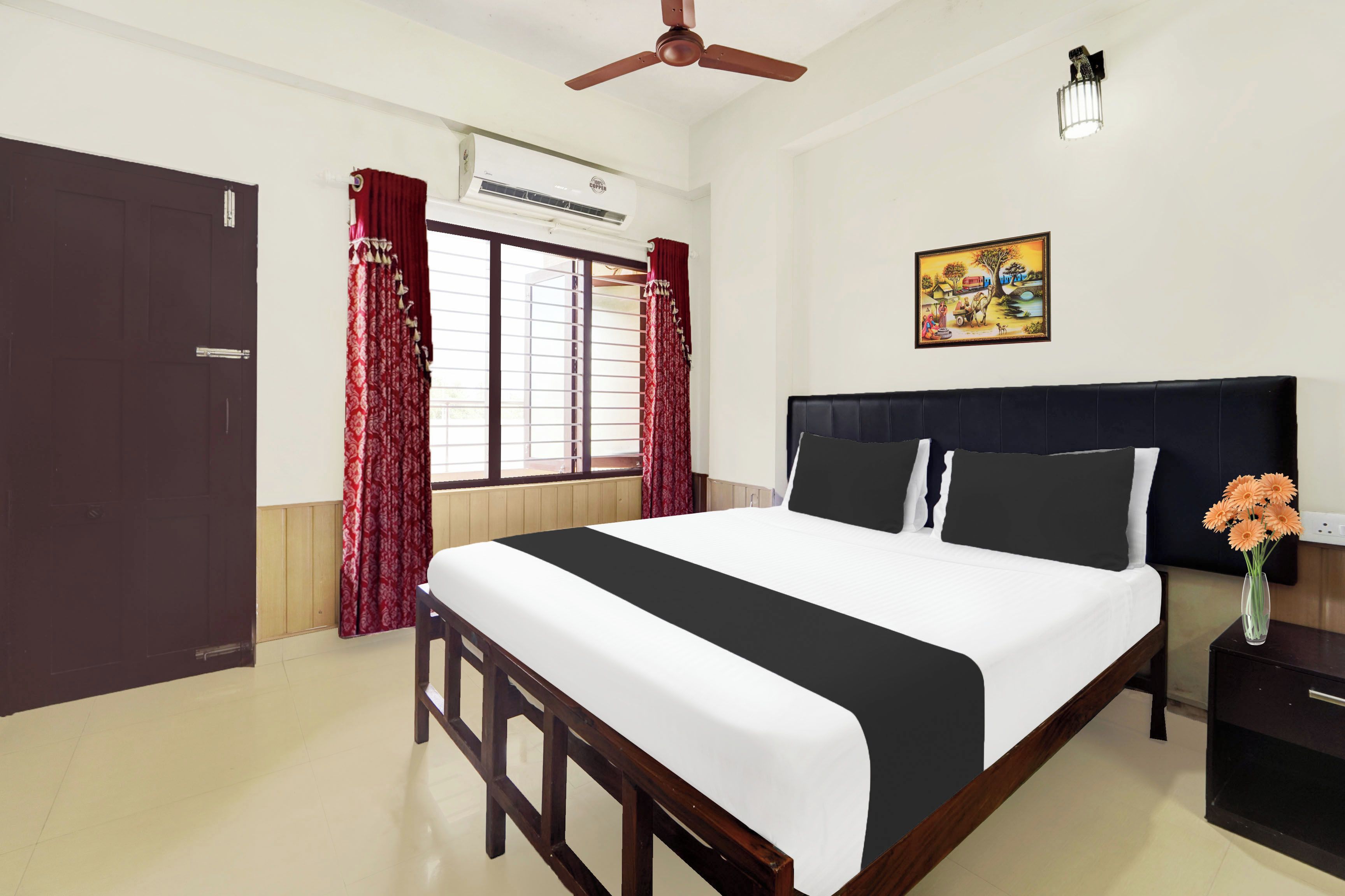 A hotel room with a bed, a ceiling fan, an air conditioning unit, a window with curtains, a nightstand with a vase of flowers, and a wall-mounted picture.