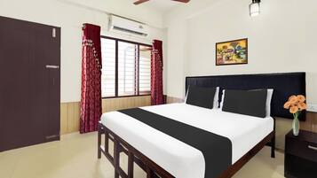A hotel room with a bed, a ceiling fan, an air conditioning unit, a window with curtains, a nightstand with a vase of flowers, and a wall-mounted picture.