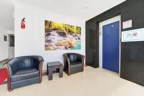 A reception area with two black leather chairs, a small table, a fire extinguisher, a blue elevator door, and a wall-mounted sign.