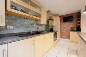 A modern kitchen with a built-in oven, a stainless steel range hood, and a flat-screen TV mounted on a brick wall. - Uncle Bill`s Cottage (Thongsbridge)